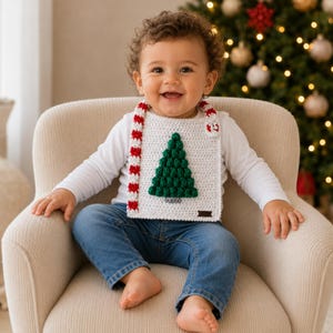 May include: A white crocheted baby bib featuring a textured green Christmas tree appliqué in the center and red and white striped trim along the edges. The bib is secured with a small white button at the top and includes a small brown rectangular tag.