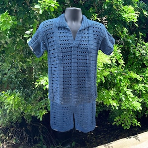 May include: A blue crochet two-piece set consisting of a short-sleeved collared shirt with a V-neckline and matching shorts. The garment features a repeating openwork stitch pattern throughout, creating a textured, semi-sheer appearance.