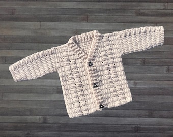 May include: A cream-colored long-sleeve crochet cardigan featuring a V-neckline and three dark, triangular buttons down the front. The garment is crafted with a textured, repeating stitch pattern throughout the sleeves and body.