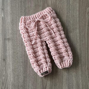 Pu&ograve; includere: Pantaloni all'uncinetto color rosa chiaro caratterizzati da un motivo a trecce testurizzato lungo le gambe. Il capo presenta un elastico in vita con cordino rosa abbinato e una piccola etichetta metallica rettangolare sull'orlo inferiore.