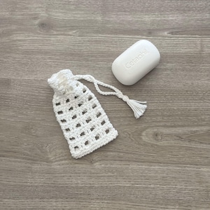 May include: White crocheted soap bag with a drawstring and tassel. The bag is sitting on a wood surface next to a white bar of Cetaphil soap.