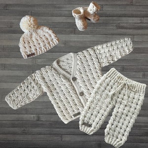 May include: A cream-colored crochet baby set consisting of a long-sleeve cardigan with three buttons, matching pants with a drawstring waist, a beanie with a pom-pom, and a pair of booties. Each piece features a textured cable knit pattern.