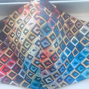 May include: A face mask with a colorful geometric pattern featuring a mix of blue, red, yellow, and gold tones. The pattern is made up of small squares and diamonds.