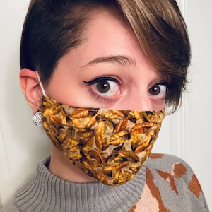 May include: A person wearing a face mask with a brown and gold leaf pattern. The mask is made of fabric and has a white elastic band. The person is wearing a gray sweater with a brown and white pattern.