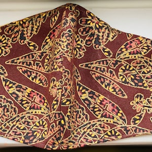 May include: A brown face mask with a floral pattern in yellow, red, and black. The mask has white elastic ear loops. The fabric features a repeating design of stylized flowers and leaves.