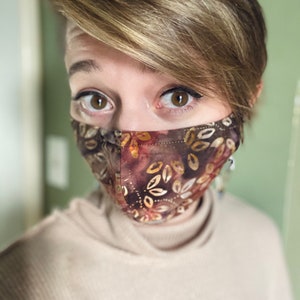 May include: A person wearing a brown and gold patterned face mask with a light brown turtleneck sweater.