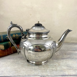 Op de afbeelding: Een zilveren theepot met een houten knop op de deksel. De theepot heeft een rond lichaam met een decoratief ontwerp en een gepolijste afwerking. De theepot staat op een houten oppervlak.