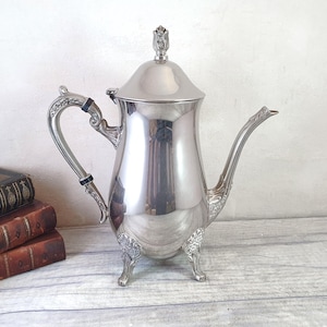 May include: A silver teapot with a floral design on the lid and handle. The teapot has three legs and a spout. The teapot is on a white wooden surface.