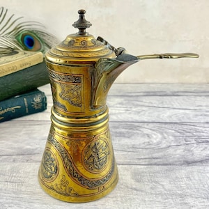 Pode incluir: Uma cafeteira de cobre com desenhos gravados intrincados. A cafeteira tem uma tampa com dobradiças e um bico com alça. A cafeteira é decorada com um acabamento dourado e prateado.