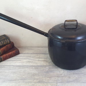 May include: A black enamel saucepan with a long, dark wooden handle and a matching lid. The lid has a small, metal handle. The pot sits on a light-coloured wooden surface, with antique books in the background.