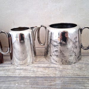 Op de afbeelding: Twee zilverkleurige kannen met handvatten. De kleinere kan heeft een tuit. Beide kannen hebben een glanzend, reflecterend oppervlak met wat slijtage. Ze staan op een houten oppervlak.