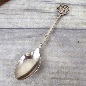 May include: A silver teaspoon with an ornate handle featuring a monogrammed design. The handle is decorative and the spoon is polished.
