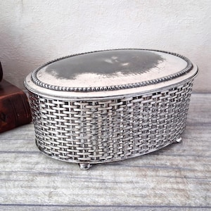 May include: Silver-toned oval jewellery box with a woven basket design. The lid is slightly domed with a beaded edge and a reflective surface. The box sits on small feet and is placed on a wooden surface.