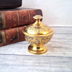 May include: A small, gold-coloured trinket box with a lid, featuring an embossed floral design. The box sits on a pedestal base. The background includes two antique books with brown covers and gold lettering.