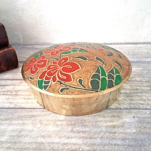 Puede incluir: Un joyero redondo de latón con un diseño floral grabado en la tapa. La superficie de la tapa está decorada con esmalte incrustado en tonos rojo vibrante y verde, sobre un fondo de latón texturizado de tono dorado.