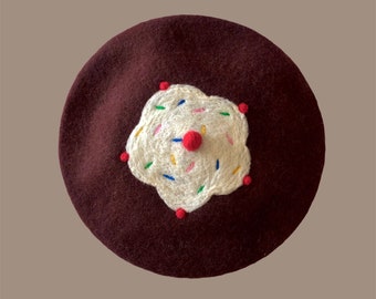 Könnte beinhalten: Eine dunkelbraune Baskenmütze mit einem aufgestickten Cupcake-Design in Weiß und Rot. Der Cupcake hat bunte Streusel.