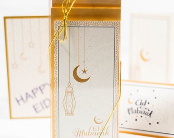 May include: A set of three gold and white greeting cards with a crescent moon and lantern design. The cards are tied together with a gold ribbon and say "Eid Mubarak".