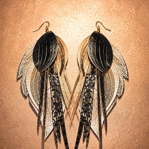 May include: A pair of layered dangle earrings shaped like wings, featuring a combination of black, gold, and metallic silver textured materials. The design includes multiple pointed, feather-like segments with intricate patterns and gold-tone hooks.