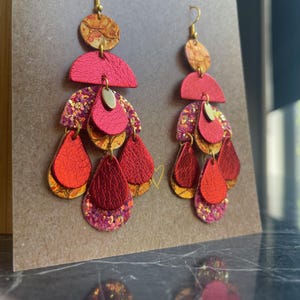 May include: A pair of long, dangling earrings with multiple layers of red and gold leather and glitter. The earrings have a teardrop shape and are attached to gold hooks.