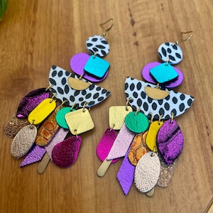 Puede incluir: Un par de pendientes llamativos y coloridos. Cada pendiente presenta un gancho dorado, un círculo con lunares blancos y negros y varias formas en morado, turquesa, dorado y rosa. Los pendientes están hechos de cuero.