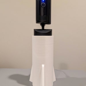 May include: A black Ring security camera mounted on a white rocket-shaped stand. The camera has a blue light on the front and the word "ring" is visible on the side.