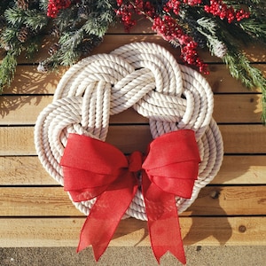 May include: A white rope wreath with a red bow. The wreath is made of thick rope and is tied in a knot design. The bow is made of a soft, red fabric and is tied in a simple knot.