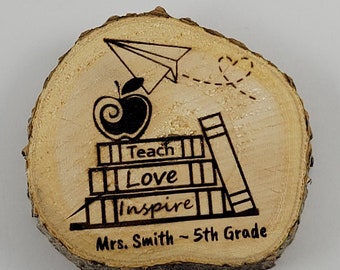 May include: A wooden round slice with a design of a stack of books, an apple, a paper airplane, and a heart. The text on the books reads "Teach Love Inspire" and the text below the books reads "Mrs. Smith - 5th Grade".