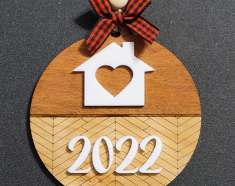 May include: A round wooden ornament with a white house cutout and a heart inside. The bottom half of the ornament has a chevron pattern and the year 2022 in white. The ornament is decorated with a red and black plaid bow.