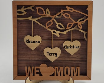 May include: Wooden framed sign with a brown wood background and a gold tree with leaves. Three hearts hang from the tree with the names Shauna, Terry, and Christine. The sign reads "WE ♥ MOM".