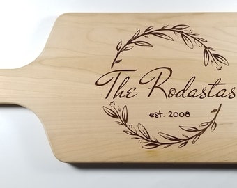 May include: A wooden cutting board with a handle, engraved with a floral wreath design and the text "The Rodastas est. 2008".