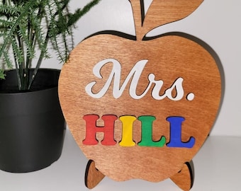May include: Wooden apple-shaped teacher desk sign with the text "Mrs. HILL" in colorful letters. The letters are red, yellow, green, and blue.