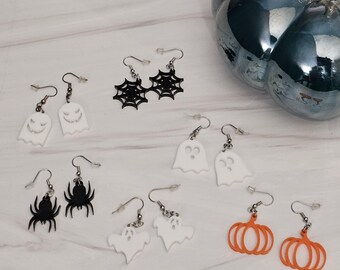 May include: A collection of Halloween-themed earrings. The earrings feature designs of ghosts, spiders, spiderwebs, and pumpkins. The earrings are in black, white, and orange. The earrings are displayed on a light-colored surface.