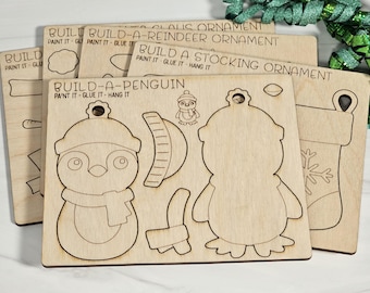 May include: Five unfinished wooden ornaments for painting and decorating. The ornaments are shaped like a penguin, reindeer, stocking, and two different styles of Christmas trees. The ornaments are labeled "Build-a-Penguin", "Build-a-Reindeer Ornament", "Build a Stocking Ornament", and "Claus Ornament".