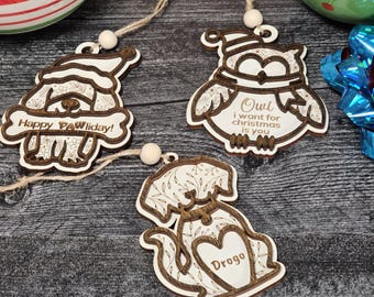 May include: Three wooden Christmas ornaments featuring a dog, an owl, and a dog. The dog ornaments have a floral design and the text "Happy Pawliday!" and "Drogo". The owl ornament has a floral design and the text "Owl I want for Christmas is you".