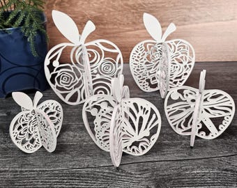 May include: Five white papercraft apples with intricate cut-out designs. The designs include flowers, swirls, and butterflies.