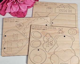 May include: Three wooden craft kits for making Valentine's Day cards. Each kit includes pre-cut pieces to assemble a heart, a teddy bear, and an envelope. The kits are labeled "Build-a-Valentine" and include instructions to paint, glue, and hang the finished creations.