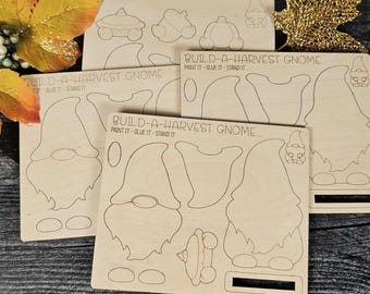 May include: Wooden craft kits with the text "BUILD-A-HARVEST GNOME" and instructions "PAINT IT - GLUE IT - STAND IT". The kits feature outlines of gnome figures and fall-themed shapes, perfect for DIY projects.