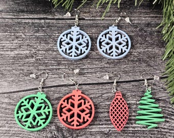May include: A collection of colorful Christmas-themed earrings. The earrings include snowflake, ornament, and Christmas tree designs in blue, green, and red. The earrings are made of wood and have silver-colored hooks.