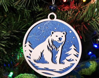 May include: A round Christmas ornament featuring a white polar bear silhouette against a blue background with a snowy scene. The ornament is made of wood and has a white outer frame. It hangs from a string with a dark blue bead.