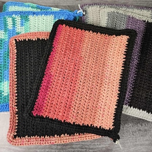 May include: A selection of handmade crochet washcloths in various colours. The washcloths have a square shape with a textured stitch pattern and a contrasting border. Colours include black, pink, orange, blue, green, and grey. The washcloths are stacked on a wooden surface.