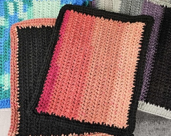 May include: A collection of handmade crochet washcloths in various colors. The washcloths feature a square shape with a textured stitch pattern and a contrasting border. Colors include black, pink, orange, blue, green, and gray. The washcloths are stacked on a wooden surface.