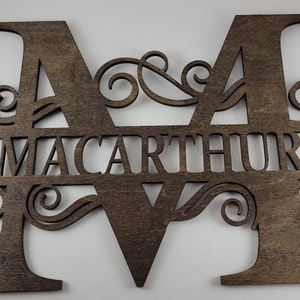 May include: A wooden sign with the last name "MacArthur" cut out in the center of the letters "M" and "M". The letters are decorated with intricate scrollwork.