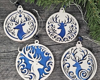 May include: Four white wooden Christmas ornaments with a deer design and blue backing. Each ornament has a different decorative pattern around the deer. The ornaments are hanging from white strings with a black bead.