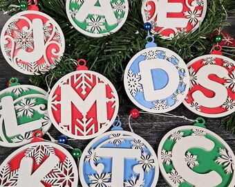 May include: A collection of round Christmas ornaments with letters, each in a different color scheme. The letters are white, with snowflake cutouts, and set against red, green, and blue backgrounds. Each ornament has a small loop for hanging.