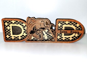 May include: Wooden sign with the word "DAD" in a decorative font. The letter "A" is replaced with a fish jumping out of the water. The sign is brown with a black and white checkered pattern. The text "Soren Bianca" is engraved on the sign.