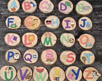 May include: A set of 26 wooden alphabet blocks, each featuring a letter of the alphabet and a colorful illustration. The blocks are painted in various colors, including red, blue, green, yellow, and orange. The illustrations include a frog, a giraffe, a house, an ice cream cone, a jellyfish, a kite, a lion, a monkey, a gear, an octopus, a penguin, a quilt, a rainbow, a sun, a tree, an umbrella, a violin, a watermelon, a xylophone, a ball of yarn, and a zebra.