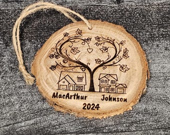 May include: A round wooden ornament with a heart-shaped tree and two houses. The tree has leaves and hearts. The houses have windows and doors. The ornament is engraved with the text "MacArthur Johnson 2024".