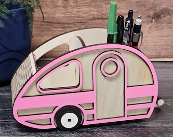May include: A pink and white wooden camper desk organizer with a slot for pens and pencils. The camper has a pink stripe and a white tire.