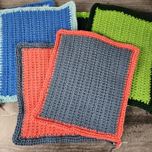 Puede incluir: Una colección de cuadrados de ganchillo hechos a mano en varios colores. Los cuadrados son en tonos de azul, coral y verde, con bordes contrastantes. El punto de ganchillo crea una superficie texturizada. Estos cuadrados podrían usarse como paños o artículos decorativos.