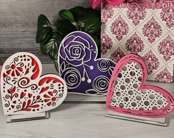 May include: Three wooden heart-shaped decorations with intricate designs. The first heart is red and white with floral patterns. The second heart is purple with a floral design. The third heart is pink with a woven pattern.